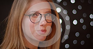 Profile shoot of blonde model in glasses with bright make-up smiles shyly and prettily on bokeh background.