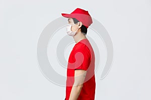 Profile shot of asian delivery guy in red cap and t-shirt, wearing protective medical mask, looking left, waiting at