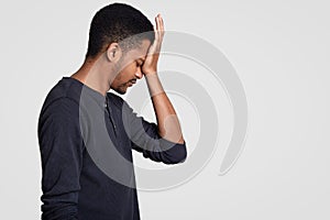 Profile shot of dark skinned man in casual outfit, hits his forehead with regret, being pessimist, feels unhappy, being in despair