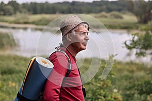 Profile shot of eldery man with backpack and blue rug, wearing red casual sweater and cap, enjoing beautifulnature near river and