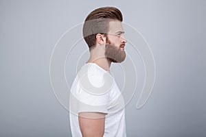 Profile shot of handsome male with trendy hairdo and beard, looks aside with serious expression, has thick red beard,