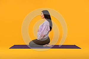 Profile shot of satisfied pregnant lady sitting on fitness mat, practicing yoga and meditation, yellow background