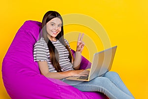 Profile side photo of young girl sit chair use laptop free-time show peace v-symbol isolated over yellow color