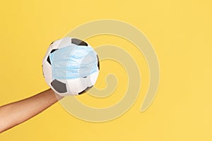 Profile side view closeup of human hand holding football or soccer ball in protective face mask, protection against coronavirus