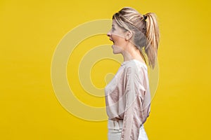 Profile side view of happy amazed blonde woman with ponytale hairstyle and in casual beige blouse, isolated on yellow background