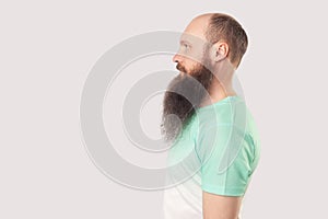 Profile side view portrait of calm relaxed middle aged bald man with long beard in light green t-shirt standing and looking