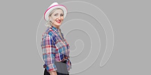 Profile side view portrait of glad satisfied successful stylish mature woman in casual style with hat standing, looking at camera