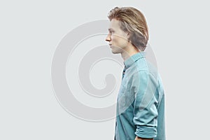 Profile side view portrait of handsome long haired blonde young man in blue casual shirt standing and looking with serious face