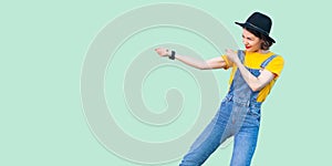 Profile side view portrait of pretty young hipster girl in blue denim overalls, yellow shirt and black hat standing with fist or