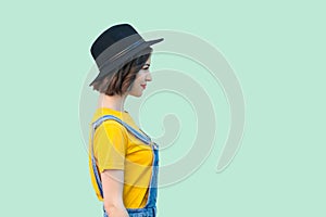 Profile side view portrait of pretty young hipster girl in blue denim overalls, yellow shirt and black hat standing, smiling and