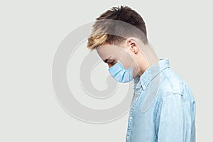 Profile side view portrait of sad upset alone young man with surgical medical mask in blue shirt standing, holding head down and