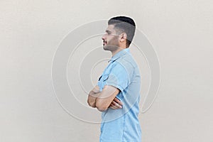 Profile side view portrait of serious calm handsome young bearded man in blue shirt standing, crossed arms and looking forward