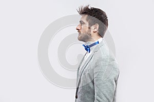 Profile side view portrait of serious handsome bearded man in casual grey suit, blue bow tie standing and looking straight wih