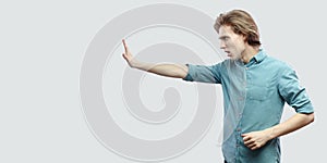 Profile side view portrait of serious handsome long haired blonde young man in blue casual shirt standing with stop hand gesture