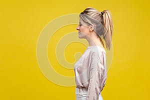 Profile side view of serious elegant young woman with ponytale hairstyle and in casual blouse,  on yellow background