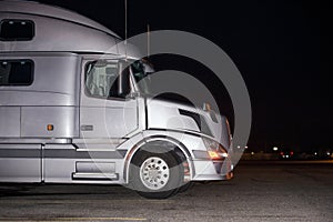 Profile of silver big rig semi truck with turned on headlight standing on dark night parking lot