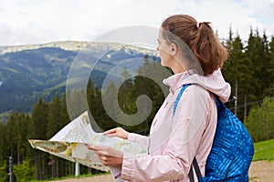 Profile of traveler woman visiting high mountins, pretty dark haired girl with map in hands and blue backpack searching right way
