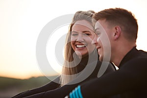 Profile View Of Couple Wearing Wetsuits On Surfing Staycation Sitting On Beach Looking Out To  Sea