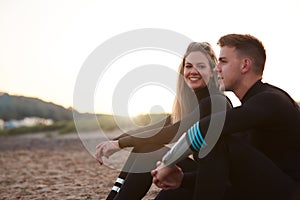 Profile View Of Couple Wearing Wetsuits On Surfing Staycation Sitting On Beach Looking Out To  Sea