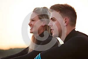 Profile View Of Couple Wearing Wetsuits On Surfing Staycation Sitting On Beach Looking Out To  Sea