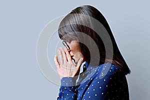 Profile view of mature sad woman, with closed eyes, folded hands, gray background