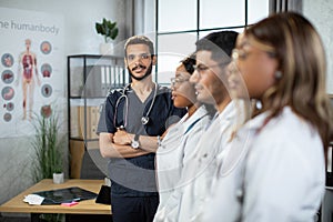 Multiracial group of medical interns in lab coats standing in a row at modern light clinic room