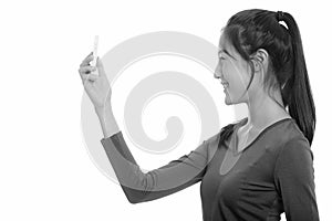 Profile view of young happy Asian teenage girl smiling while taking selfie with mobile phone
