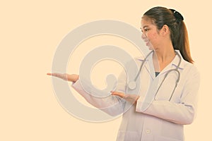 Profile view of young happy Asian woman doctor smiling while showing something