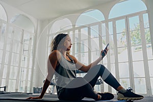 Profile view of young multiracial woman in sportswear sitting and using mobile phone at the gym