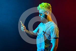 Profile of young adult bearded hipster guy with green hair wearing denim overalls, holding smart phone in hands, looks at display