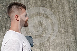 Profile of young good looking bearded guy standing outdoors against grey modern loft modern loft wall with calm face emotion.