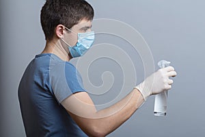 Profile of young man in surgical mask holding up bottle of disinfectant spray on a gray studio background, fight against spread of