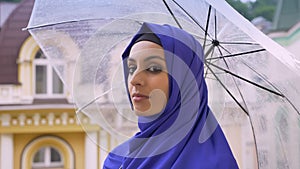 Profile of young muslim woman in hijab holding umbrella during rain, turning and looking at camera