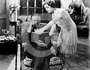 Profile of a young woman showing a book to a man sitting in an armchair