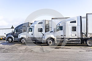 Profiles of big rigs semi trucks with semi trailers stand on wide parking lot of truck stop