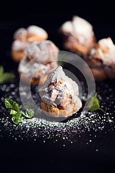 Profiteroles or cream puff with filling, falling powder sugar topping. Berries, mint on black background