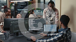 Programer sitting at desk with multiple screens running code talking with colleague