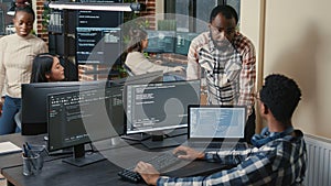 Programer sitting at desk with multiple screens running code talking with colleague