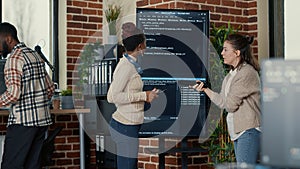 Programmer holding digital tablet analyzing code on wall screen tv while colleague is looking for errors