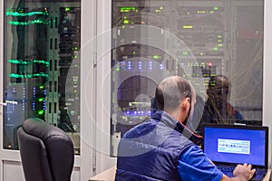 A programmer  with a laptop sits in front of the glass door of the server room. The  system administrator works in a modern data