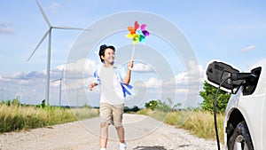 Progressive young boy playing windmill toy next to EV car at wind turbine farm.