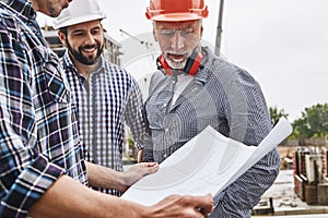 Project discussing. Senior engineer in formal wear and helmet looking at construction drawing with two young cheerful