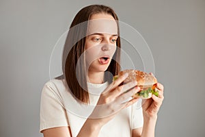 Proper nutrition, healthy fast food, unhealthy choice. Young amazed astonished woman wearing white t-shirt holding in hand burger