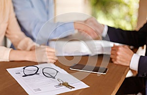 Property agreement and house key on table with real estate agent and clients shaking hands on background, closeup
