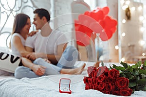 Proposal ring and red roses on the bed with couple in love on background.Man makes present to his lovely sweetheart girl.