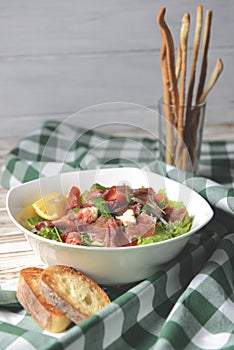 Prosciutto and arugula salad in a white bowl over wooden background. Green plaid tablecloth