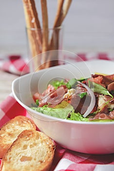 Prosciutto and arugula salad in a white bowl over wooden background. Red plaid tablecloth