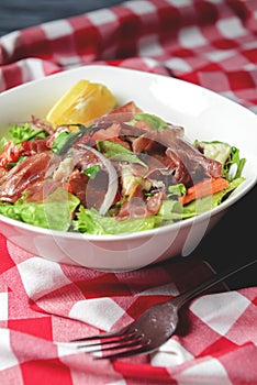 Prosciutto and arugula salad in a white bowl over wooden background. Red plaid tablecloth