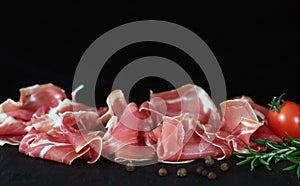 Prosciutto crudo or jamon meat with rosemary on serving board on black background. Sliced into thin pieces of ham