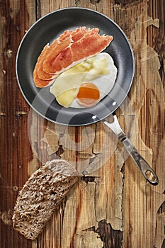 Prosciutto Rashers With Fried Egg In Frying Pan With Slice Of Brown Bread Set On Old Cracked Wooden Garden Table photo